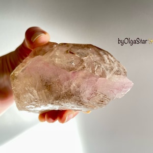 May include: A raw, translucent crystal displaying a mix of brown and pink tones. The crystal is held in a hand, highlighting its natural, uneven form. The image includes the text "byOlgaStar".