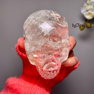 Pode incluir: Uma caveira de cristal clara e esculpida com traços faciais detalhados. A caveira é segurada em uma mão com uma manga vermelha difusa. O fundo é cinza sólido com algumas flores brancas.