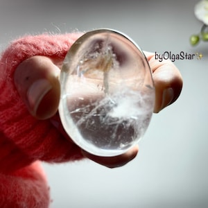 Può includere: Un cristallo trasparente di forma ovale con inclusioni interne, tenuto in una manica rosa. Il cristallo ha un aspetto nuvoloso e piumoso all'interno, con linee e imperfezioni visibili. L'immagine è ben illuminata, evidenziando la trasparenza e la consistenza del cristallo. Il testo "byOlgaStar" è visibile.
