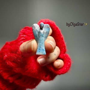 May include: A small, blue and grey angel figurine is held in a red knitted glove. The angel has outstretched wings and a detailed carved design. The background is a soft grey, with the text "byOlgaStar" visible.
