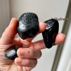 May include: A hand holding several polished, black tourmaline stones. The stones have a smooth, reflective surface and vary in size and shape. The image is well-lit, highlighting the natural texture and color of the stones.
