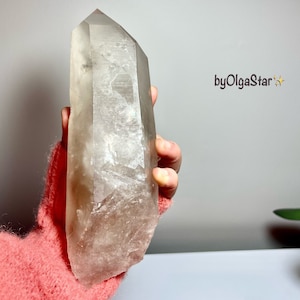 Può includere: Un grande cristallo di quarzo trasparente con una punta. Il cristallo è tenuto in mano da una persona ed è di colore marrone chiaro. Il cristallo ha una consistenza naturale e ruvida ed è un oggetto decorativo.