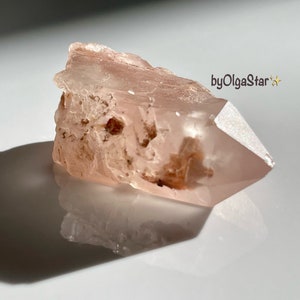 Puede incluir: Un cristal de cuarzo rosa en bruto con una punta. El cristal tiene una superficie rugosa y texturizada y un aspecto translúcido, con áreas de inclusiones rosas y marrones más oscuras. La imagen incluye el texto "byOlgaStar".