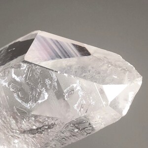 Può includere: Primo piano di una punta di cristallo di quarzo trasparente. Il cristallo ha una punta sfaccettata e un aspetto traslucido, con striature e inclusioni interne visibili. La forma generale è una forma geometrica naturale.