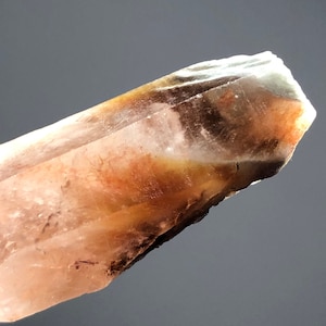 Può includere: Primo piano di un cristallo traslucido con una punta affilata. Il cristallo presenta un gradiente di colori, tra cui sfumature di arancione, marrone e bianco. La superficie sembra avere imperfezioni e texture naturali, suggerendo che si tratta di un esemplare grezzo e non lucidato.