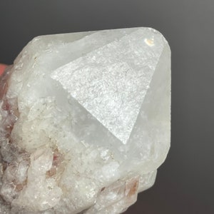 May include: Close-up of a clear quartz crystal with a pyramidal tip. The crystal has a rough, textured surface and a pale, translucent appearance. The base of the crystal has smaller, reddish-brown crystal formations.