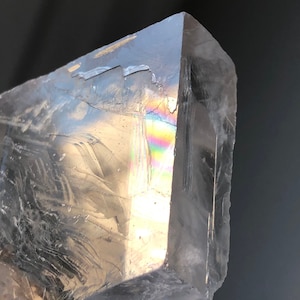 Può includere: Primo piano di un cristallo trasparente con una punta. Il cristallo ha una superficie fratturata e mostra uno spettro di colori arcobaleno dove la luce lo attraversa. Il cristallo è posto su uno sfondo scuro.