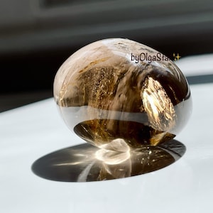 Può includere: Cristallo trasparente e arrotondato con inclusioni dorate e marroni interne. La superficie lucida riflette la luce, creando un effetto scintillante. Il cristallo proietta un'ombra su una superficie bianca. Il testo "byOlgaStar" è visibile.
