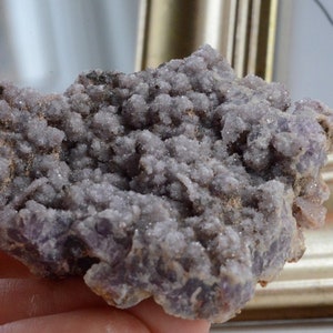May include: A close-up of a cluster of amethyst crystals. The crystals are a soft purple color with a textured, bumpy surface. The cluster sits on a brown base, with a gold-colored frame in the background.
