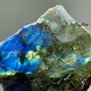Puede incluir: Primer plano de una piedra preciosa de labradorita con una vibrante exhibición de iridiscencia azul, verde y dorada. Los bordes ásperos y naturales de la piedra son visibles, y la superficie es texturizada. El texto "byOlgaStar" está en la esquina superior derecha.
