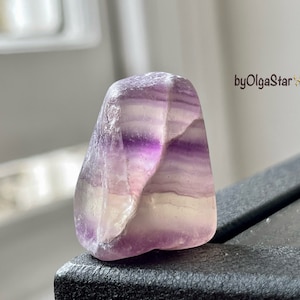 Peut inclure: Une pierre de fluorite polie avec des bandes de teintes violettes, blanches et claires. La pierre a une forme lisse et arrondie et un aspect translucide. Le texte "byOlgaStar" est visible dans le coin supérieur droit.