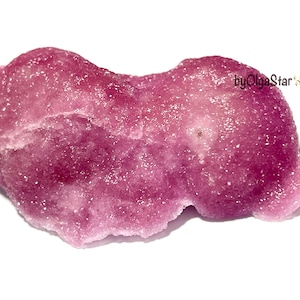 May include: A raw, irregularly shaped magenta crystal cluster with a sparkling, granular surface texture. The stone features deep pink and translucent violet hues throughout its crystalline structure. The text byOlgaStar is visible in black.