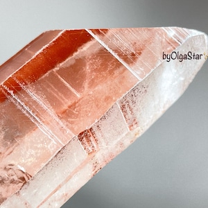 May include: Close-up of a crystal with a reddish-orange hue. The crystal has a sharp, pointed tip and a faceted surface. The text "byOlgaStar" is visible on the side of the crystal.