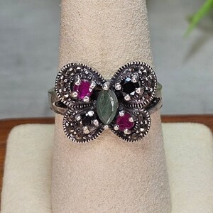 May include: Silver-toned ring with a butterfly design. The ring features a central green stone, surrounded by red and black gemstones, and accented with small, silver-colored beads. The ring is displayed on a white, textured surface.