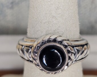 May include: A silver ring featuring a circular black gemstone. The ring has a braided pattern around the stone and on the band. The ring is presented on a white surface.