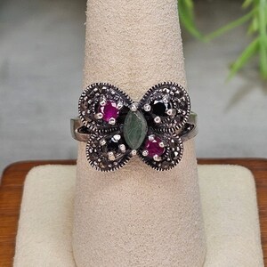 May include: A silver butterfly ring with marcasite detailing. The ring has a central green stone, with black and red gemstones on either side, forming a symmetrical pattern. The ring is shown on a neutral-toned ring stand.