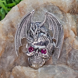 Puede incluir: Colgante de calavera plateada con diseño de dragón. La calavera tiene ojos color rubí y está adornada con un dragón y alas. El colgante se exhibe sobre una superficie de piedra clara.
