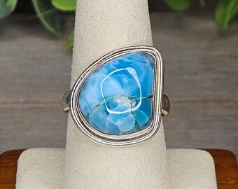 May include: A silver ring with a large, light blue gemstone with white marbling. The stone is set in a silver bezel. The ring is displayed on a white stand. The gemstone's shape is irregular. The ring is on a white jewellery stand.