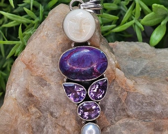 May include: A silver pendant featuring a white carved moon face, a large oval purple stone, amethyst teardrop and oval stones, and a pearl. The pendant is displayed on a light brown rock with green foliage in the background.