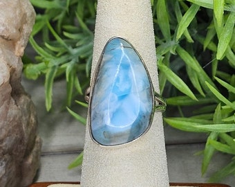 May include: A silver ring with a large, pear-shaped larimar gemstone. The stone is light blue with white marbling, set in a simple silver band. The ring is displayed on a white, textured surface.