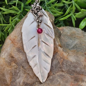 Puede incluir: Un colgante nacarado en forma de hoja con detalles grabados, acentuado por un diseño floral plateado y una pequeña gema roja redonda. El colgante se muestra sobre un fondo de piedra natural con follaje verde.