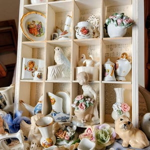 Może przedstawiać: Biała drewniana gablota wypełniona różnymi małymi figurkami porcelanowymi. Kolekcja obejmuje wazony, łabędzie, anioły i kompozycje kwiatowe. Tło przedstawia nuty.