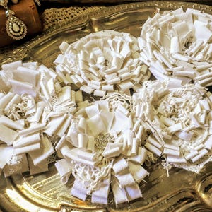 Può includere: Cinque fiori in tessuto bianco disposti su un vassoio d'argento. Ogni fiore è realizzato con strati di nastri bianchi e strisce di pizzo, con un centro decorato da strass trasparenti. I petali sono piegati e arricciati per creare una forma circolare.