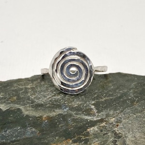 May include: A silver ring featuring a hammered spiral design. The ring is displayed on a grey rock.