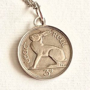May include: A silver-coloured circular coin pendant on a delicate chain. The coin features a raised relief of a hare in the centre, with the text 'leat' on the left, 'Reul' on the right, and '3d' at the bottom, all enclosed within a beaded border.