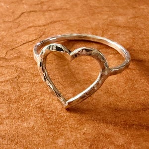 May include: A silver ring featuring a central open heart shape. The band and the heart outline have a distinct hammered, textured finish that catches the light. The metal has a bright, reflective silver tone throughout the entire piece.