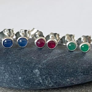 May include: A set of six silver stud earrings with different coloured gemstones. The earrings feature blue, red and green gemstones.