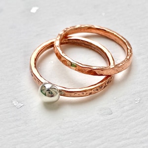May include: Two hammered copper rings with a silver bead on one ring. The rings are stacked on top of each other.