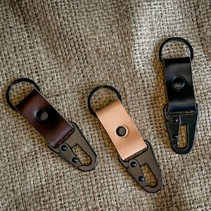 Puede incluir: Tres llaveros de cuero con ganchos de metal negro. Los llaveros son marrón, beige y negro.