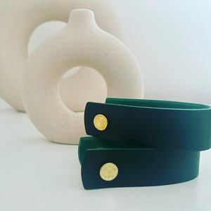 May include: Two dark green leather bracelets with gold metal accents. The bracelets are stacked on top of each other.