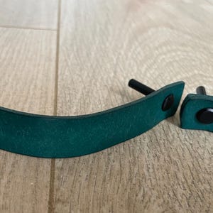 Puede incluir: Dos tiradores de armario de cuero verde azulado con herrajes negros. Uno es un mango largo y curvo, y el otro es un tirador más pequeño en forma de lazo. Ambos tienen remaches y postes negros para el montaje. Sobre una superficie de madera clara.