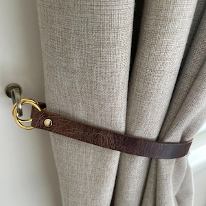 May include: A brown leather curtain tieback with a gold-coloured ring and a brass-coloured screw. The tieback is wrapped around a beige curtain. The curtain is hanging on a brass-coloured rod.