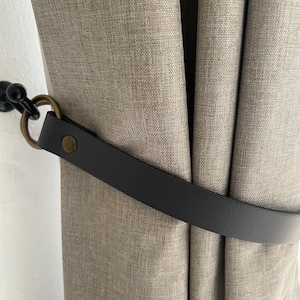 May include: A close-up of a beige curtain with a black leather tieback. The tieback is secured to a metal hook on the wall.
