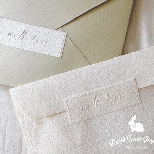 May include: Two white envelopes with a textured surface. The envelopes have a rectangular label with the text "with love" embossed on them. The bottom envelope also has the text "Rabbit Loves Graphics melbourne" printed on it.