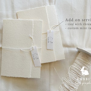 May include: Two white folded cards with a frayed edge, tied together with twine and a small tag that says "C+B 27-03-2021". A spool of white twine is in the background with the text "RABBIT love graphics".