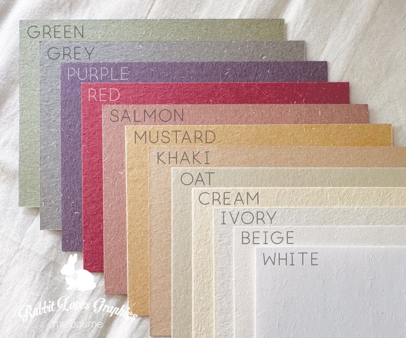 May include: A collection of 12 paper samples in various colors, including green, grey, purple, red, salmon, mustard, khaki, oat, cream, ivory, beige, and white. The paper samples are arranged in a fan shape, with the darkest color at the top and the lightest color at the bottom. The paper samples are all textured and have a natural look.