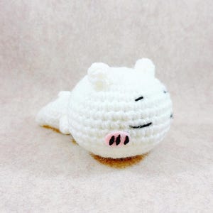 May include: A handmade white crochet pig with black eyes and a pink snout. The pig is lying down, with its body and head visible. The crochet animal is a cute, whimsical design.