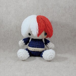 May include: A handmade crochet doll featuring a character with white and red hair, blue eyes, and a navy blue outfit. The doll is sitting with its legs extended and has a beige trim around the waist. The doll is made of yarn.