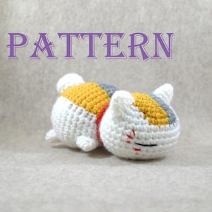 May include: Crocheted white, yellow, and gray cat with red accents. The cat is lying on its side with its head resting on its paws. The cat has a bell around its neck and a coin on its forehead. The cat is a traditional Japanese lucky charm.