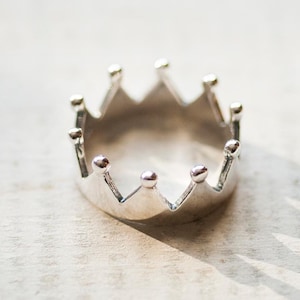 May include: A silver crown ring. The ring features a series of pointed arches, each with a small, round detail. The ring is displayed on a light-toned surface, with a shadow cast to the side.