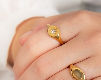 May include: Three gold rings on a hand. The first ring is a simple oval band. The second ring is a signet ring with a star and a small diamond. The third ring is a band with multiple small stones.