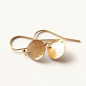 May include: A pair of gold-toned drop earrings featuring small, circular, hammered metal discs suspended from curved ear wires. The surface of each disc has a textured, reflective finish that catches the light, creating a bright, metallic appearance.