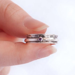May include: A silver ring with a small purple gemstone set in the center. The ring is made of three bands that are stacked on top of each other.