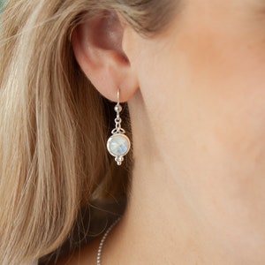 May include: Silver dangle earrings featuring a round moonstone gemstone. The earrings have a delicate design with small beads.
