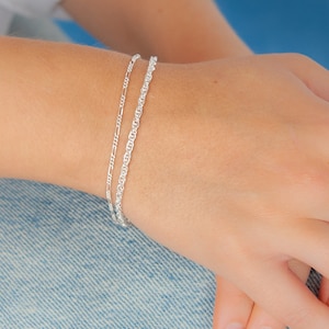Könnte beinhalten: Zwei silberne Kettenarmbänder am Handgelenk einer Person. Die Armbänder sind übereinander gelegt, wobei eine dünnere Kette auf einer dickeren Kette liegt.