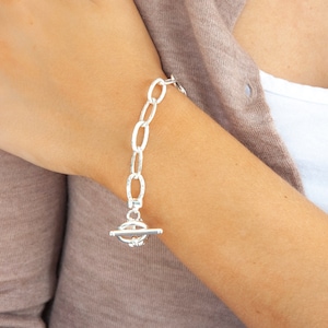 May include: A silver-colored bracelet with an oval link chain and a toggle clasp. The bracelet is worn on a wrist, with a neutral-toned top and cardigan in the background.
